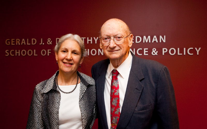 Alice Lichtenstein and the late Stan Gershoff pose in front of a red wall bearing the friedman school's full name. 